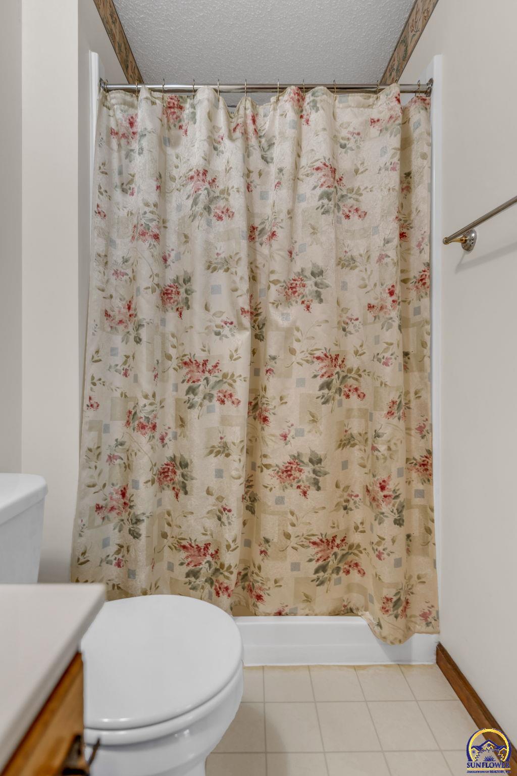 Bathroom with a cream floral shower curtain, white toilet, and pale tiled floor.