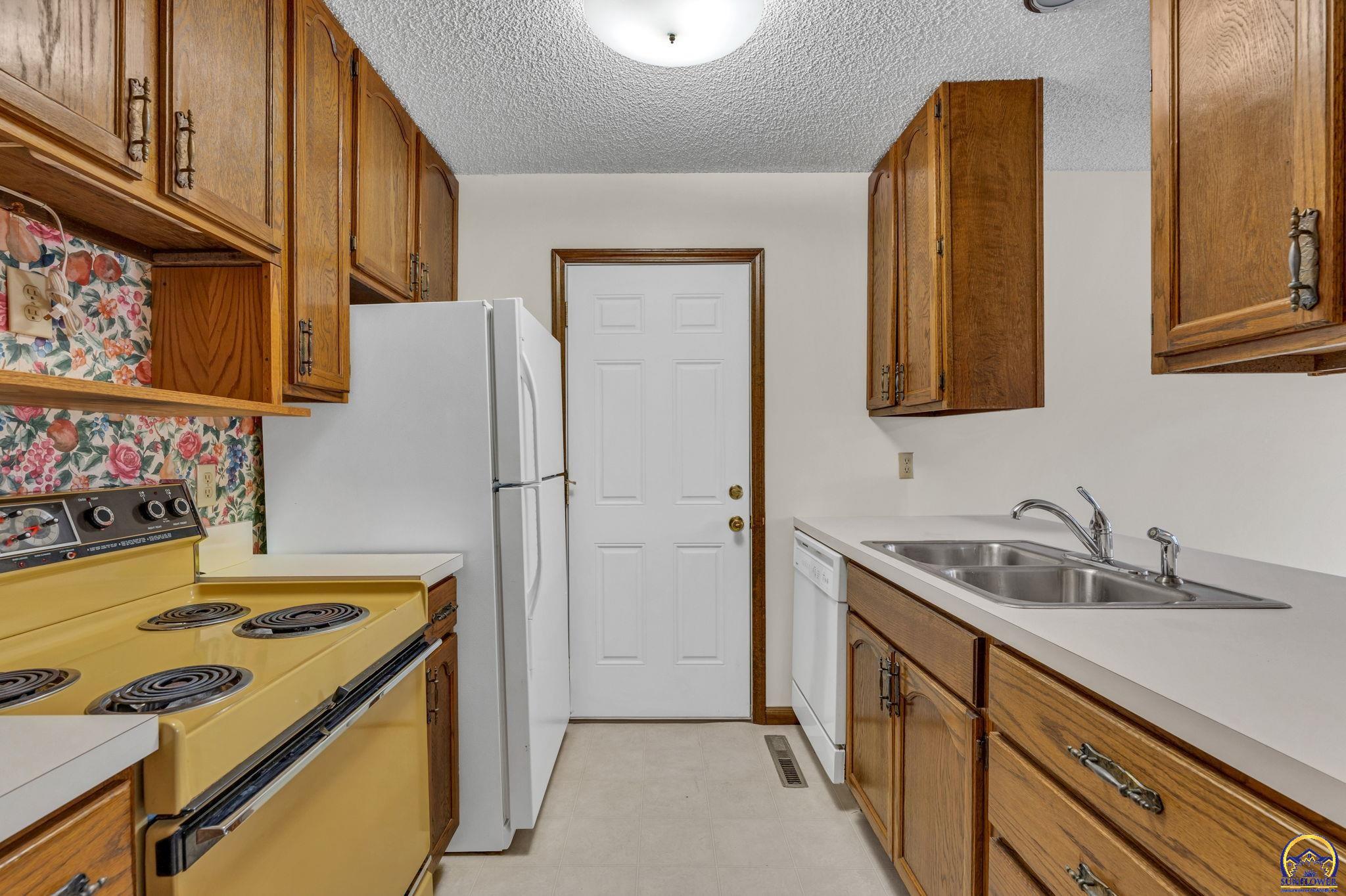 Kitchen with wood cabinets, floral wallpaper, yellow range, white refrigerator, and stainless steel double sink.