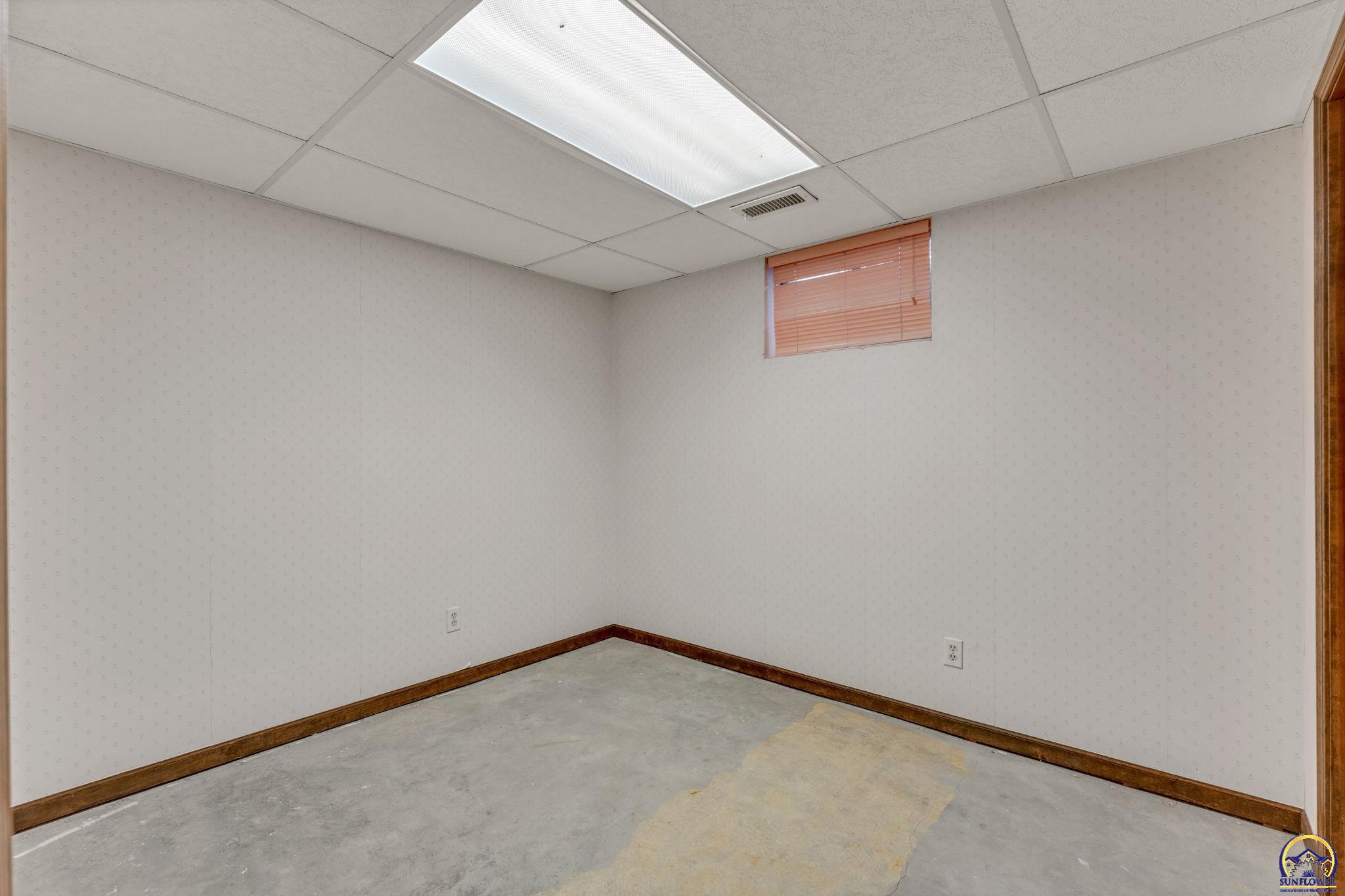 Empty basement room with a concrete floor, small window with blinds, and suspended ceiling.