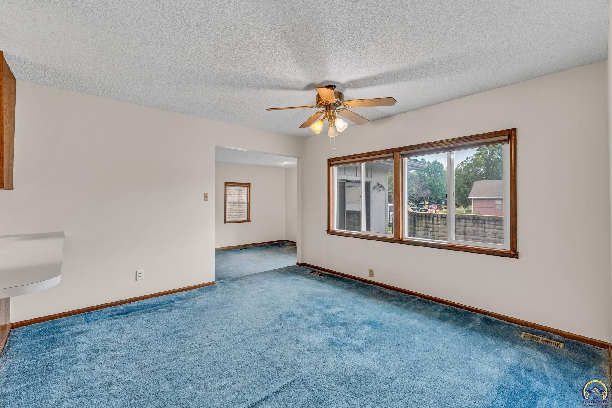 Empty living room with blue carpet, a ceiling fan, and a large wood-trimmed window.