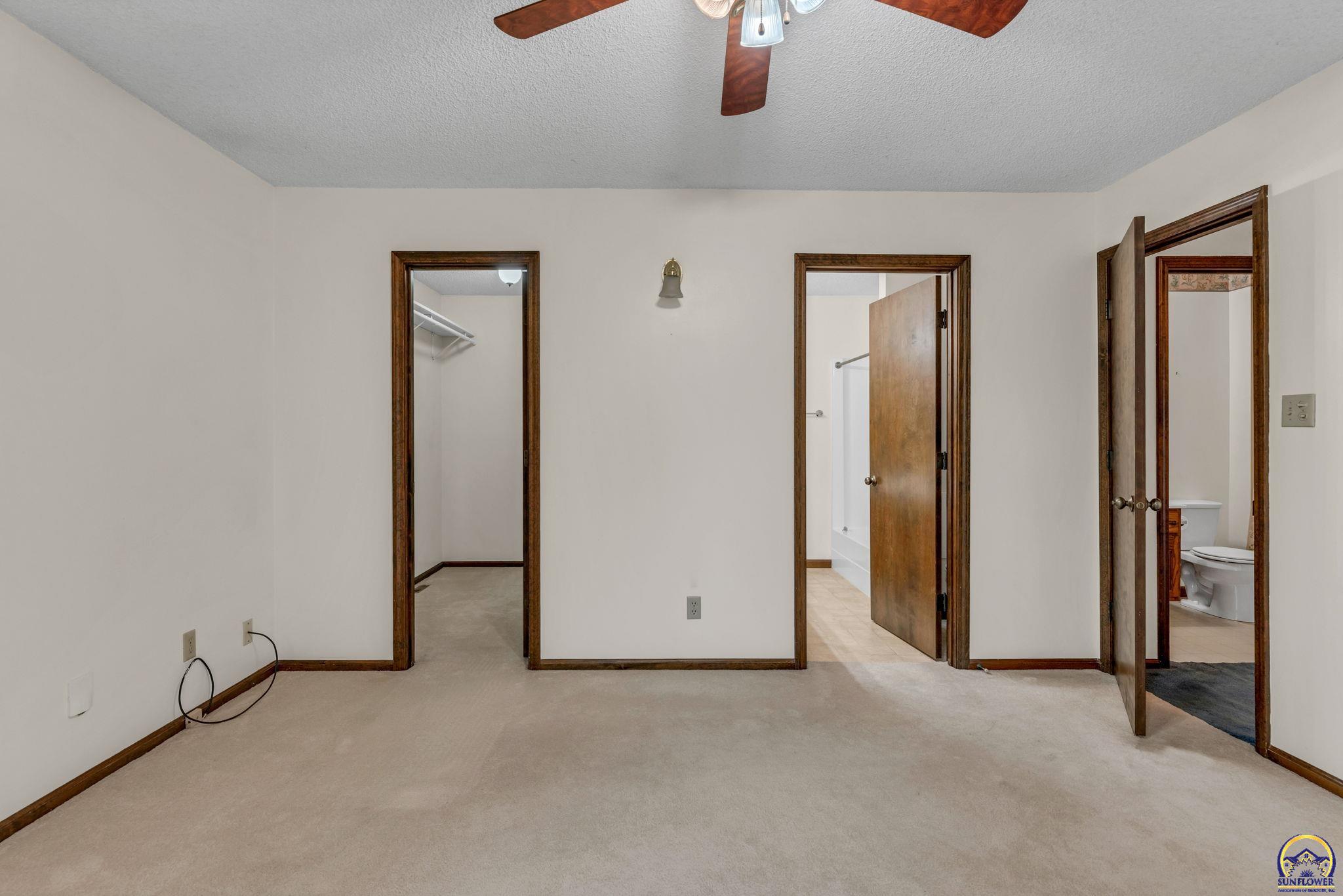 Empty carpeted bedroom with a ceiling fan and doorways to a closet and bathrooms.