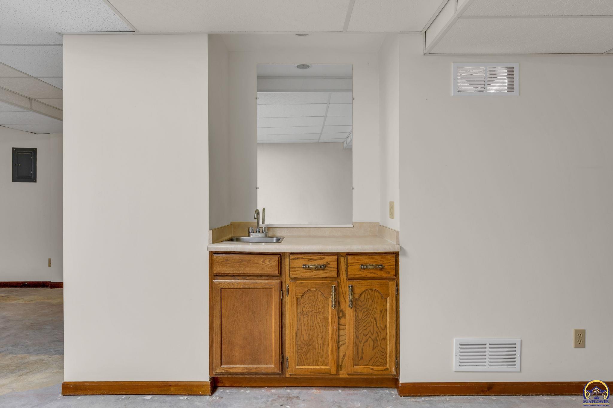 Small sink and wooden cabinets beneath a large mirror in a vacant interior.
