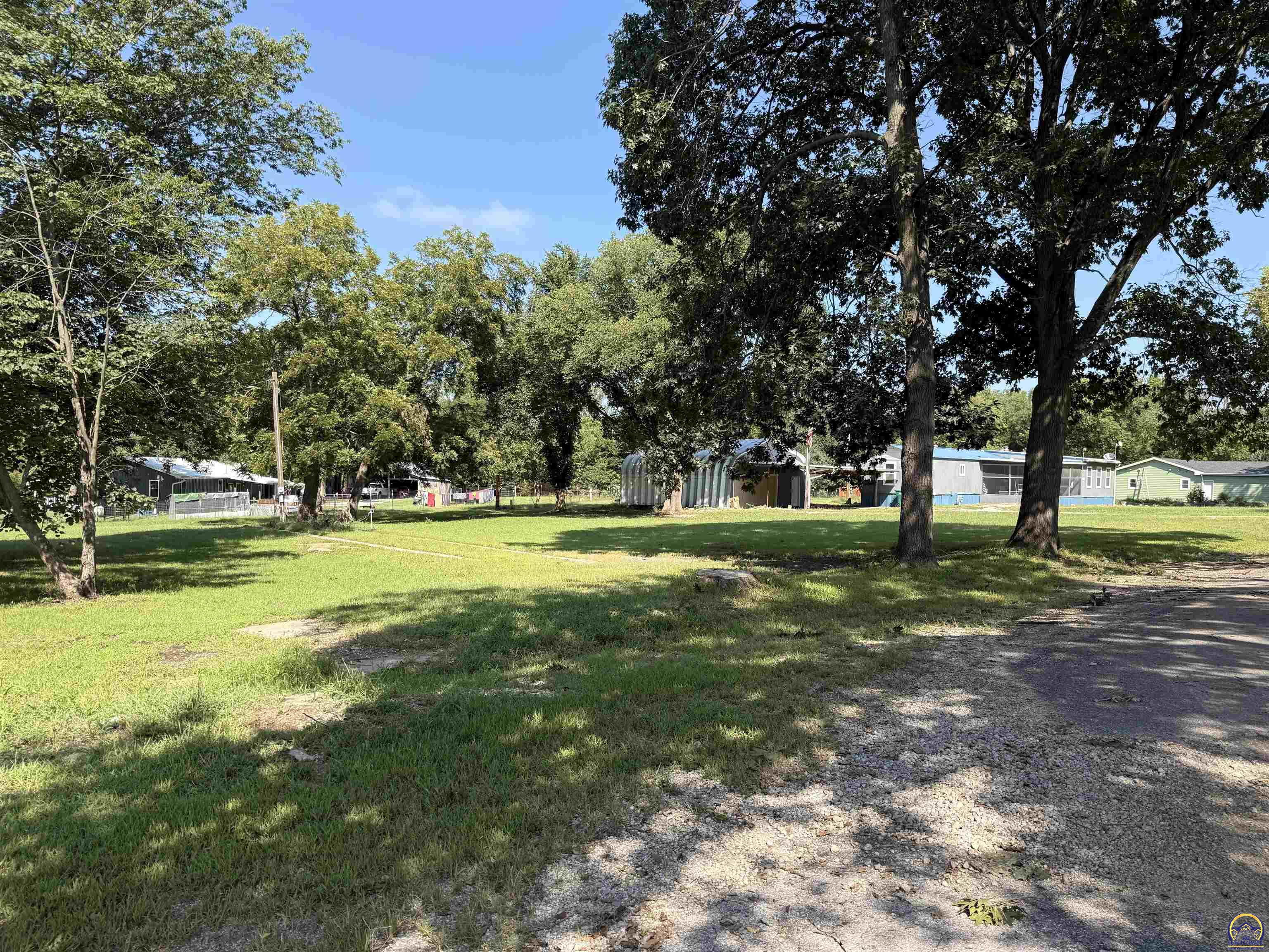 Tree-shaded grassy property with a gravel drive and several small buildings in the background.