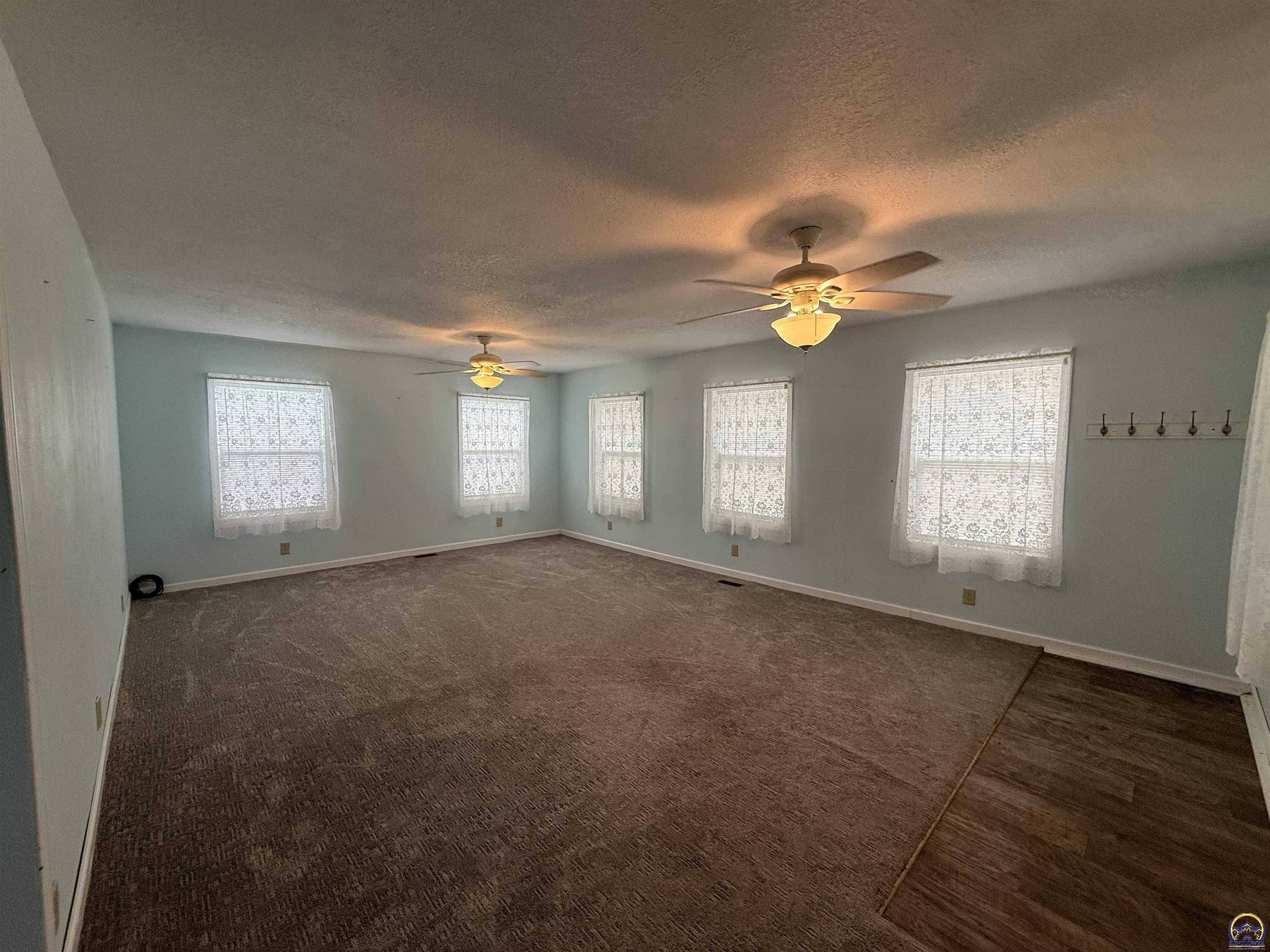 Vacant living room with five curtained windows, brown carpet, and two ceiling fans.
