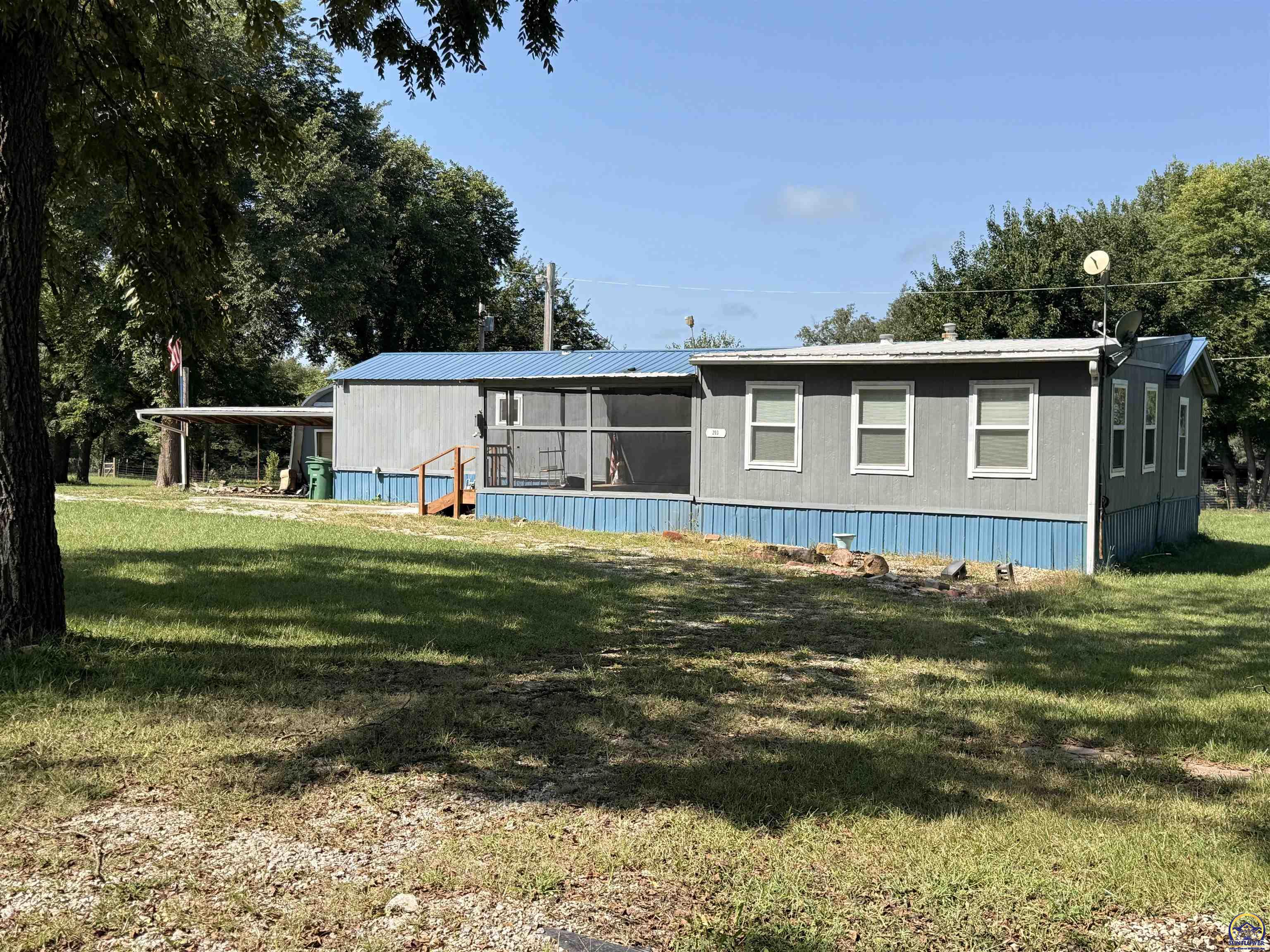 Gray manufactured home with blue skirting and screened porch on a shaded grassy lot.