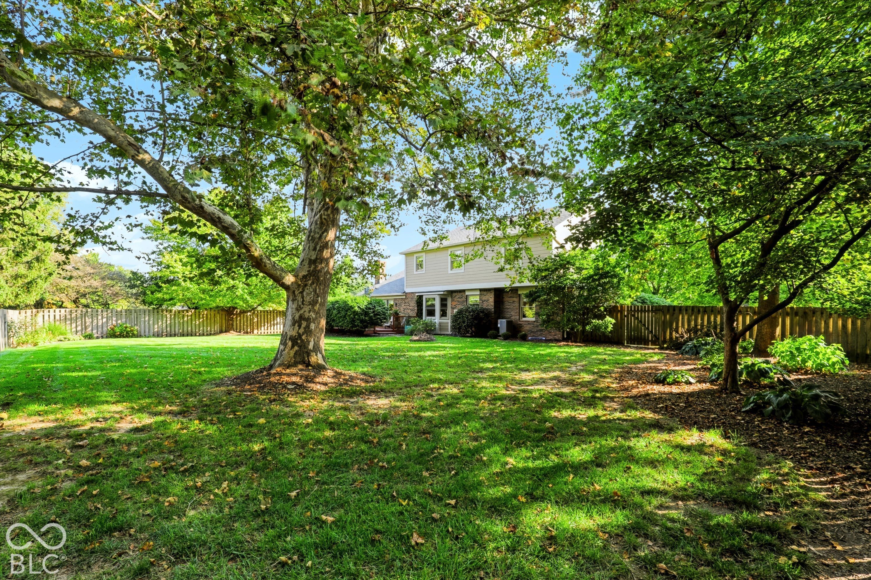 Two-story home beyond a fenced backyard lawn, shaded by mature trees.