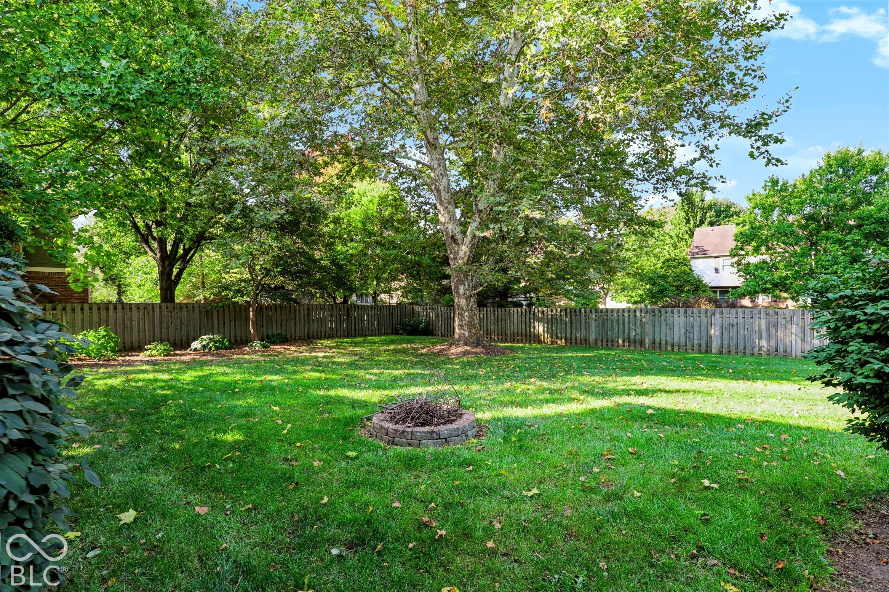 Shaded backyard with a large lawn, mature trees, wooden fence, and circular stone fire pit.