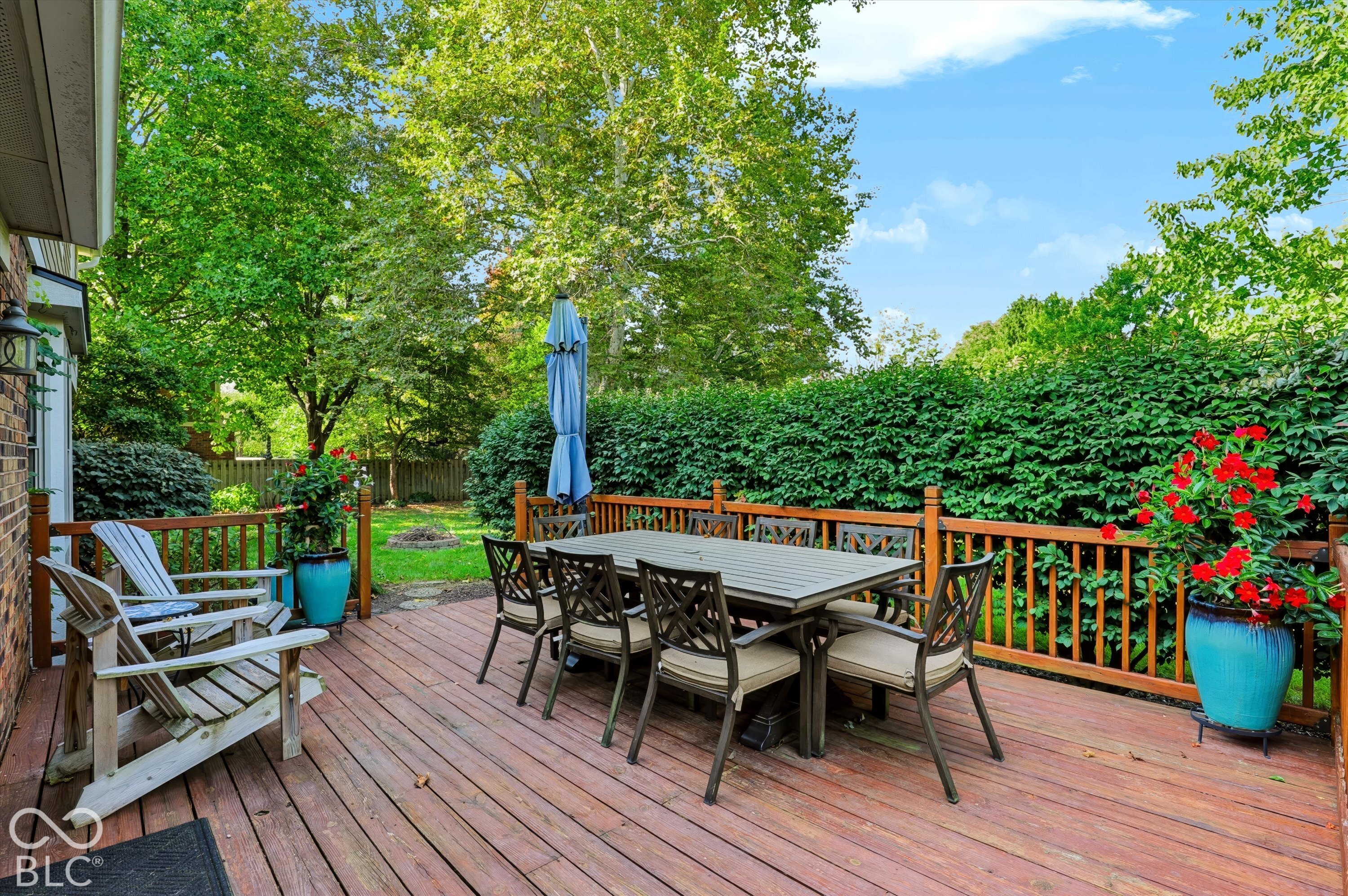Backyard deck with outdoor dining furniture, lounge chairs, and flowering planters beside tall hedges.