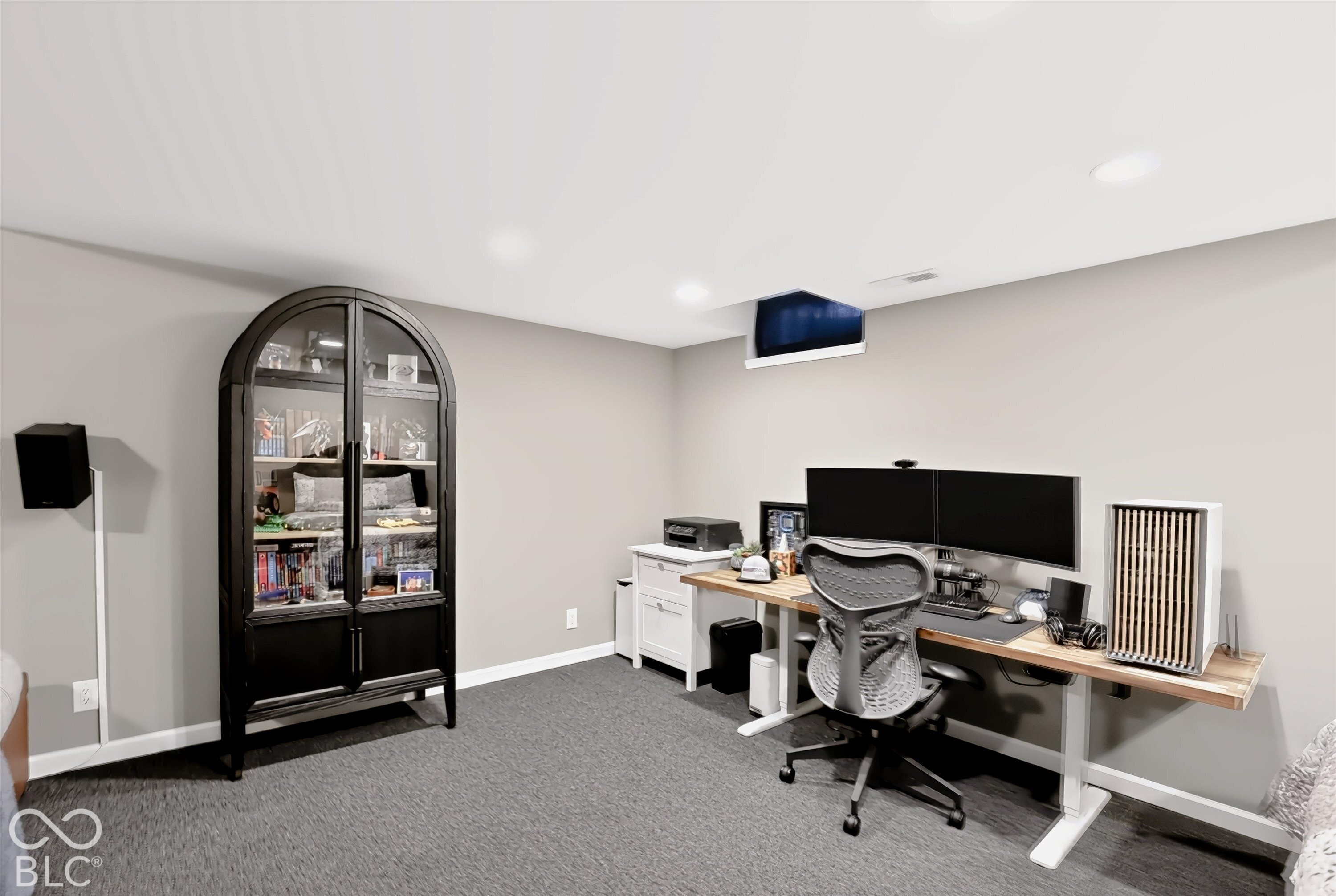 Home office with dual monitors, a wood desk, ergonomic chair, and tall glass-front cabinet.
