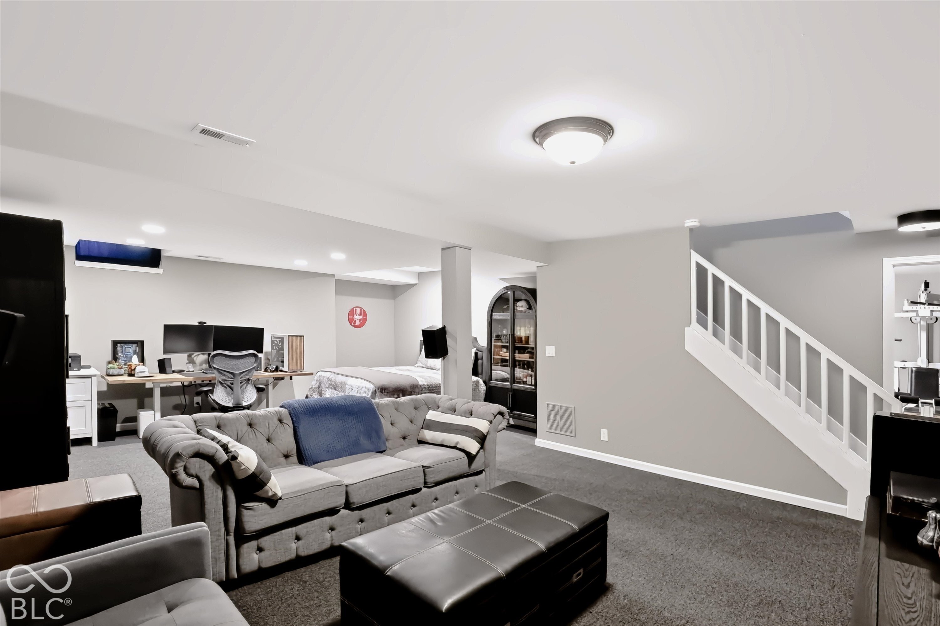 Furnished basement living area with a gray sofa, workstation, bed, and staircase.