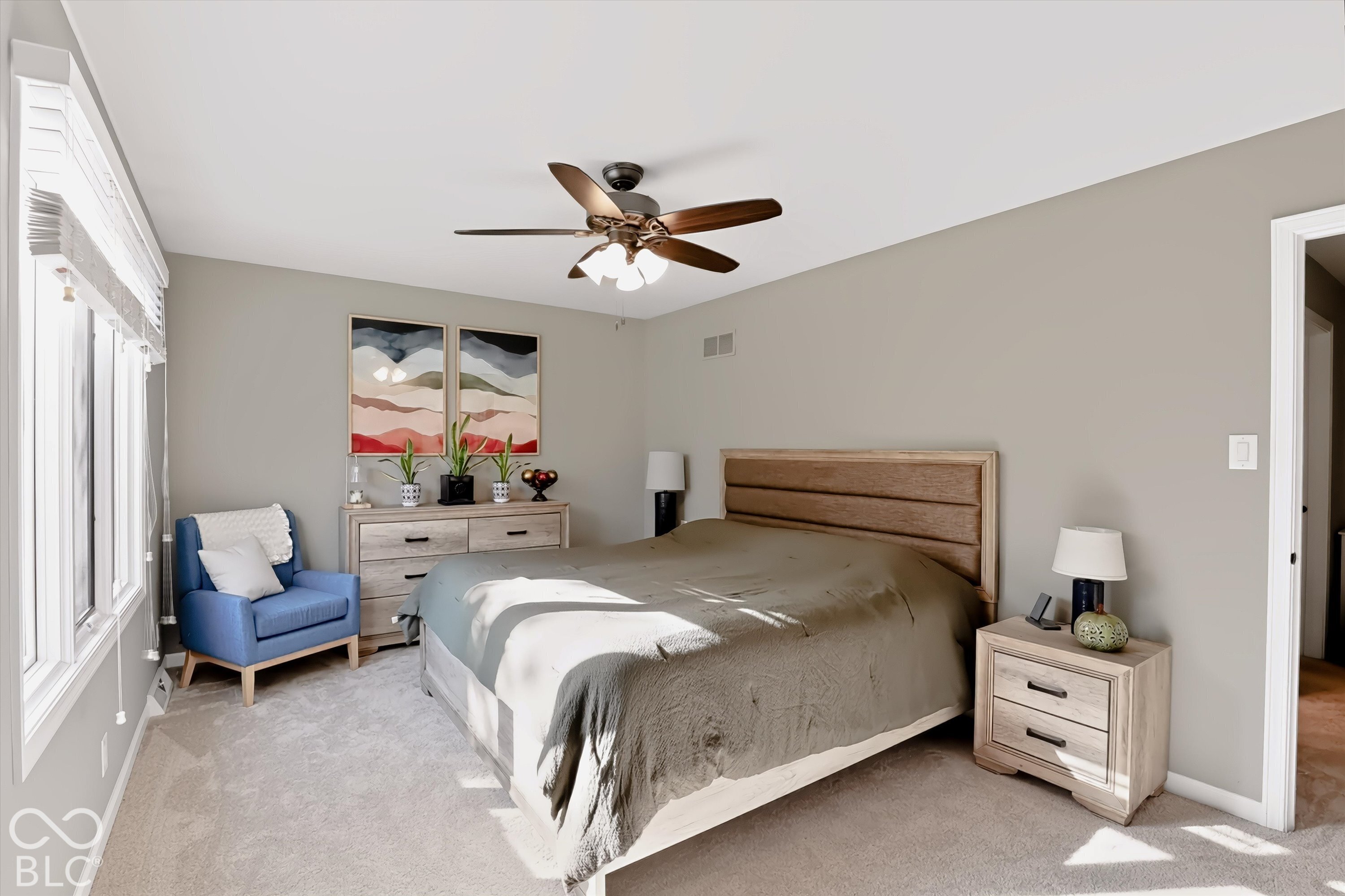 Furnished bedroom with a wooden bed, blue armchair, matching light-wood furniture, and bright windows.
