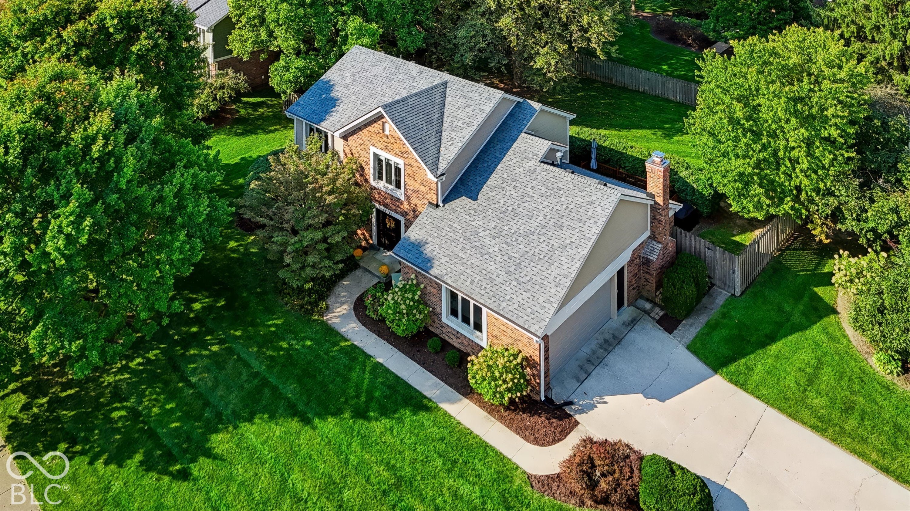 Aerial view of a brick house with a gray roof, broad driveway, landscaped lawn, and fenced backyard.