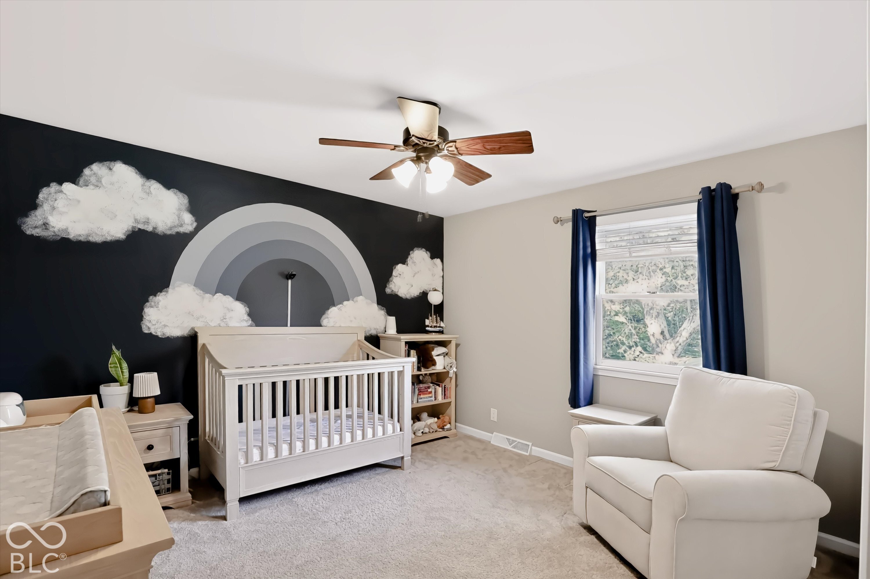 Nursery with a white crib beneath a gray rainbow-and-cloud mural and a white upholstered glider by the window.