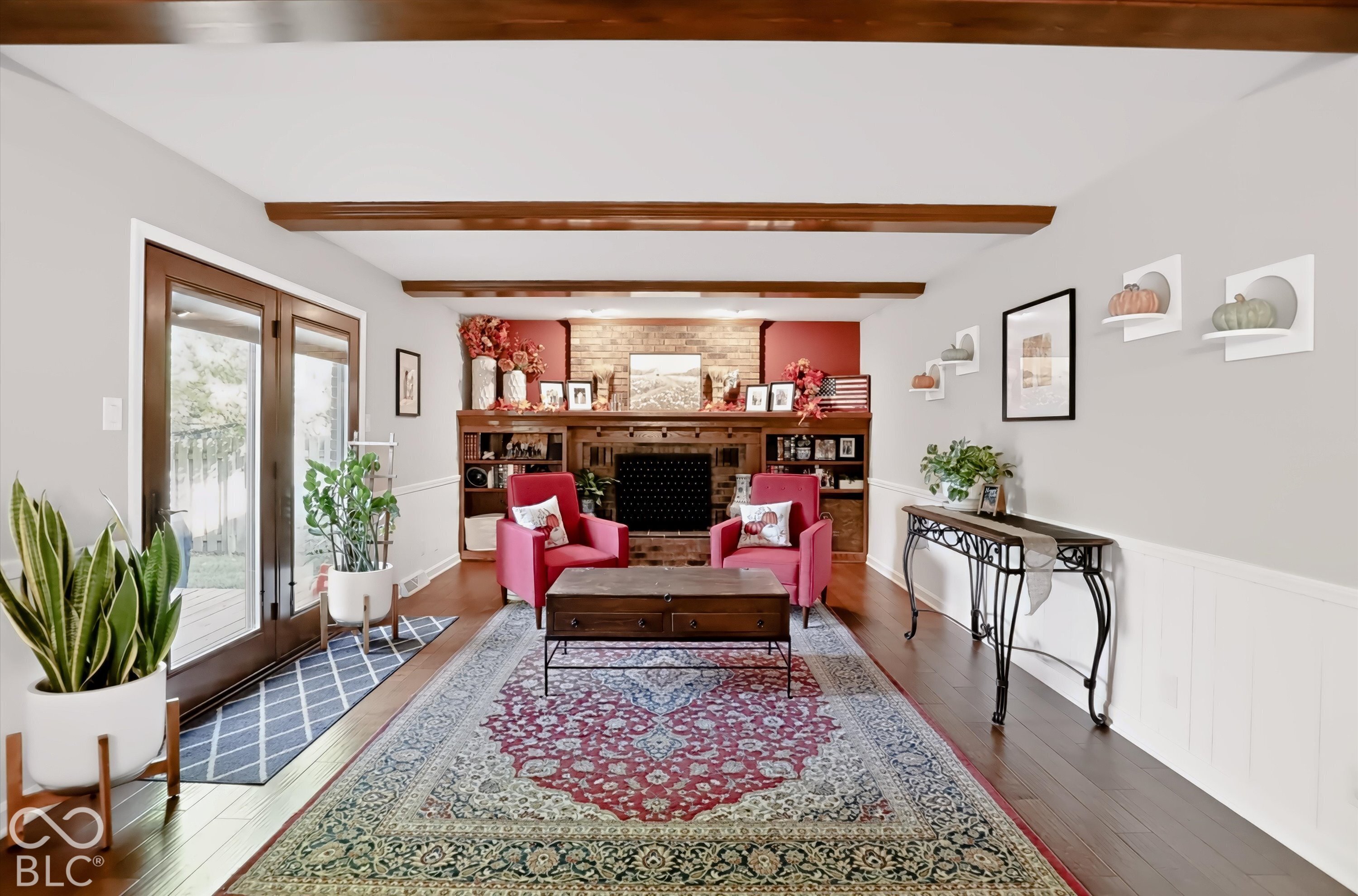 Living room with a brick fireplace, built-in shelving, red armchairs, and dark wood ceiling beams.