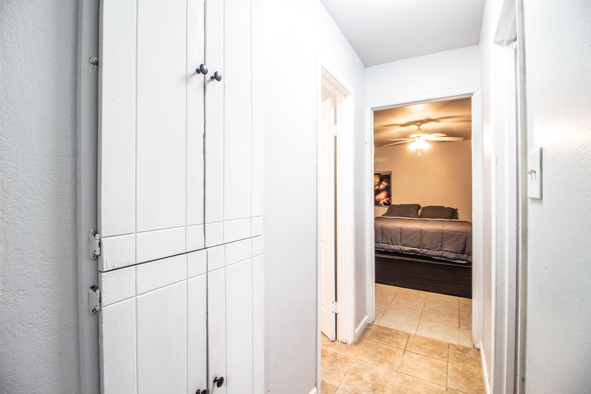Tiled hallway with white cabinets opening onto a bedroom with a bed and ceiling fan.