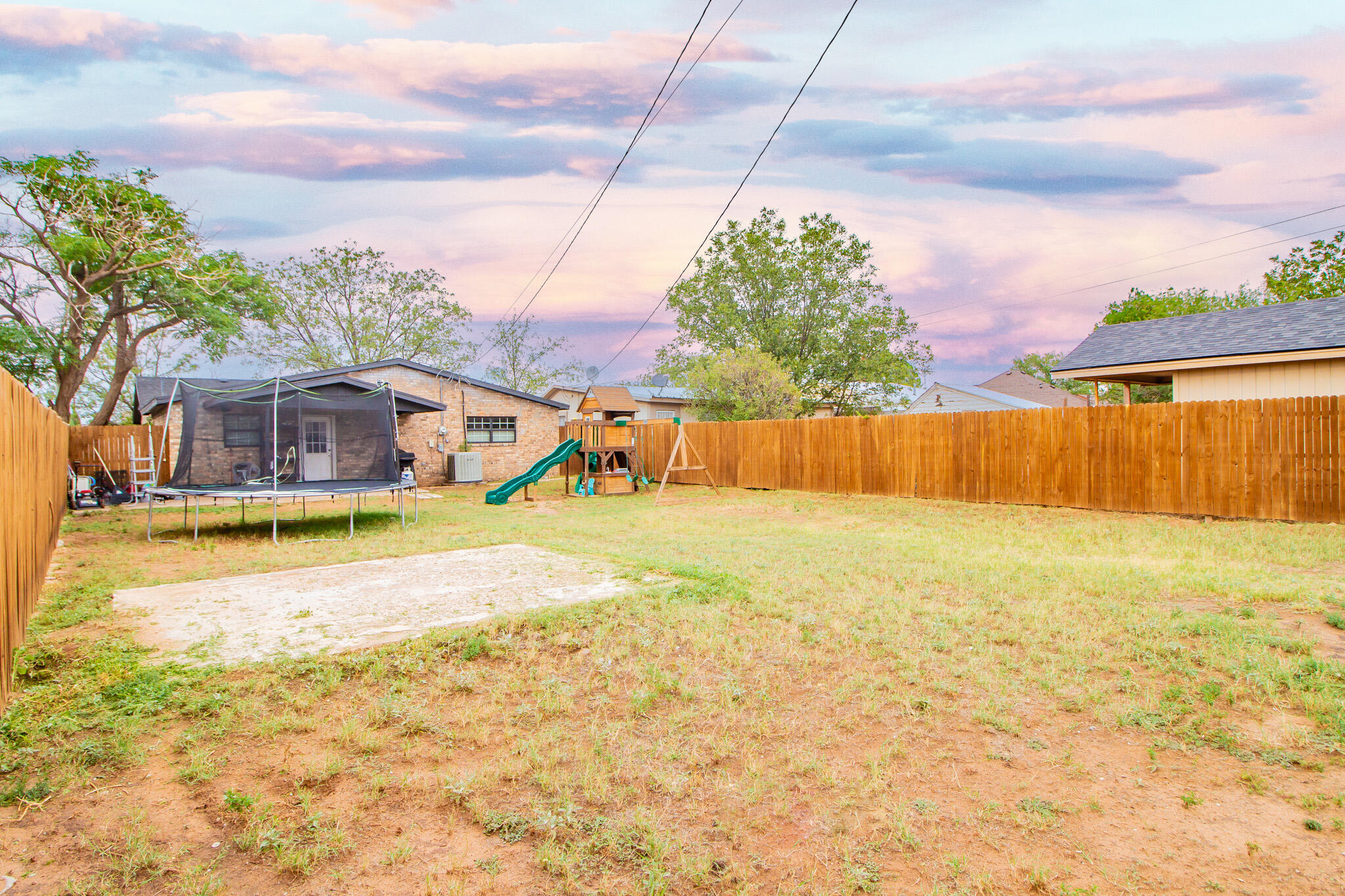 Fenced backyard with a trampoline, children’s playset, patchy lawn, and concrete patio.
