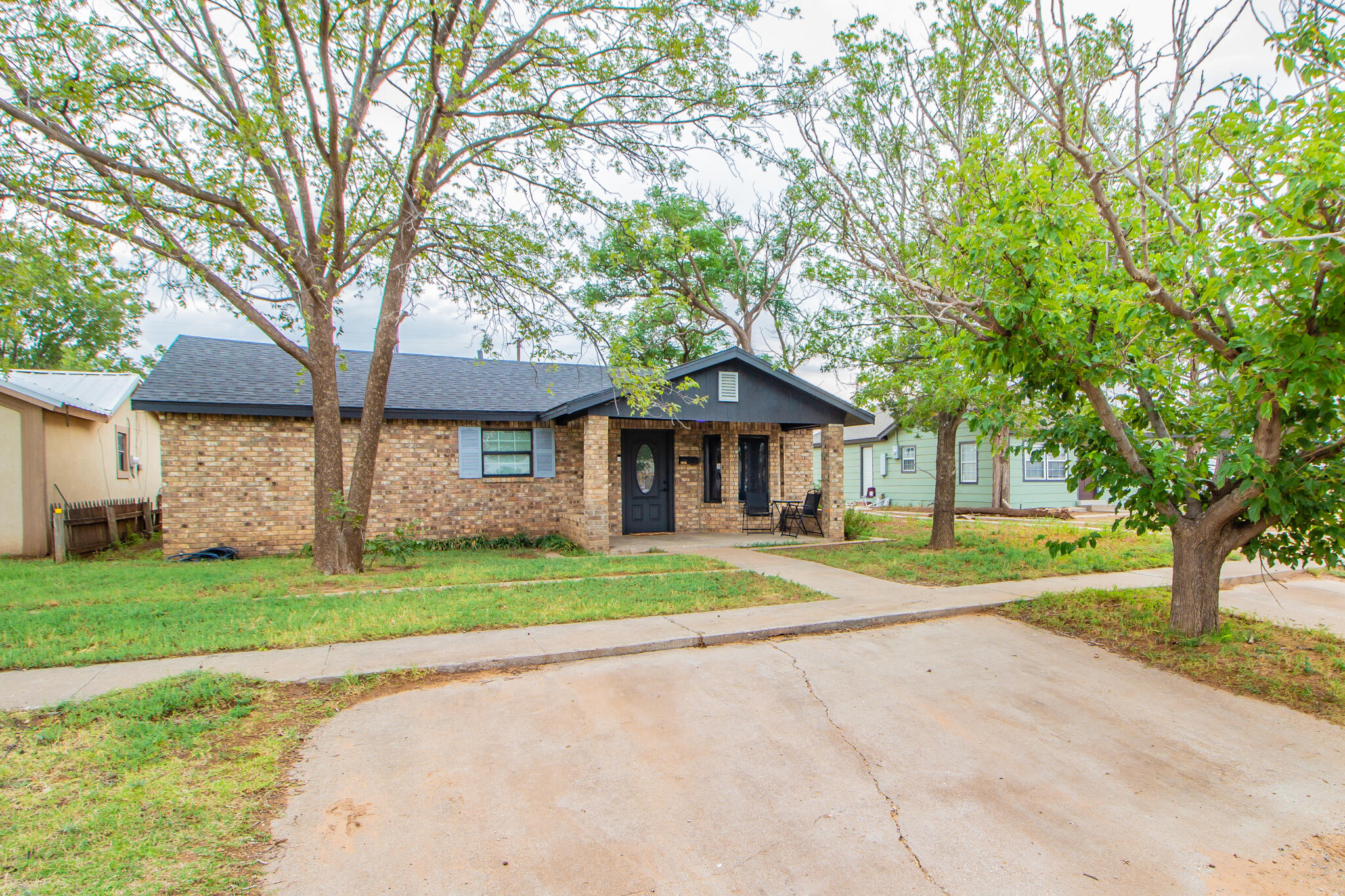Single-story brick house with a covered porch, mature shade trees, and a wide concrete driveway.