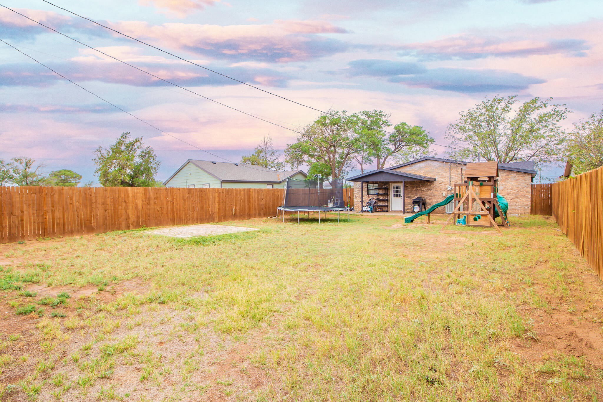 Fenced backyard with a trampoline and wooden playset beside a brick outbuilding.