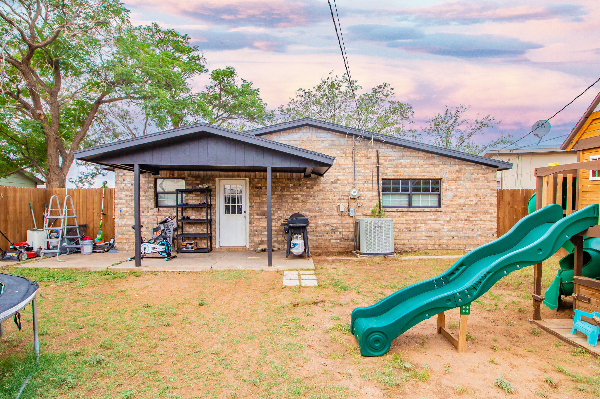 Brick house backyard with covered patio, barbecue grill, and large green playset slide.
