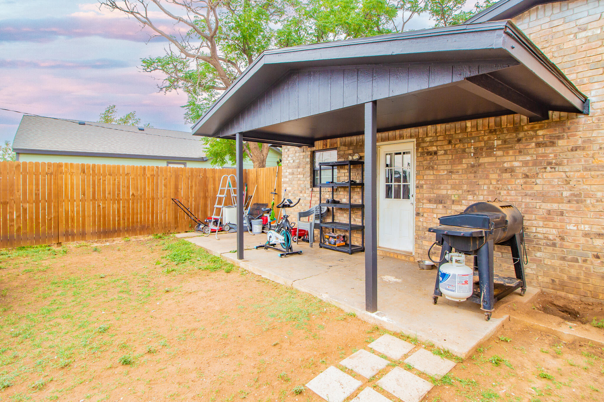 Covered concrete backyard patio beside a brick house, with a wooden fence and barbecue grill.