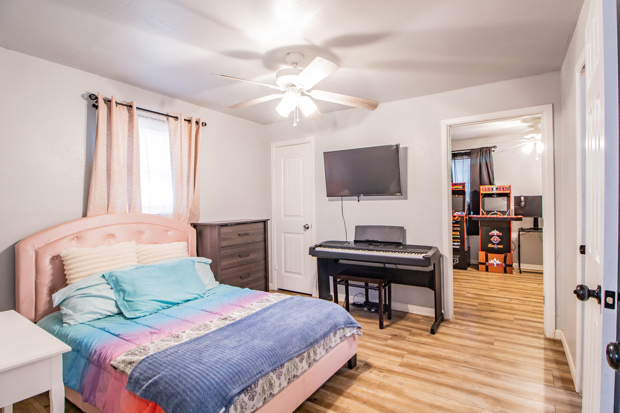 Bedroom with a pink bed, blue bedding, digital piano, and arcade machines visible through the doorway.