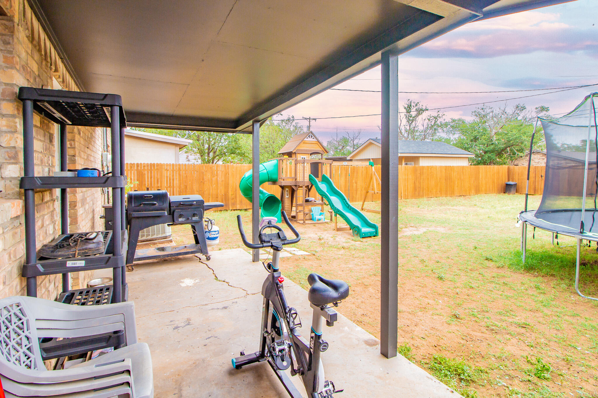 Covered patio overlooking a fenced yard with a playset and trampoline; grill and exercise bike sit under the roof.