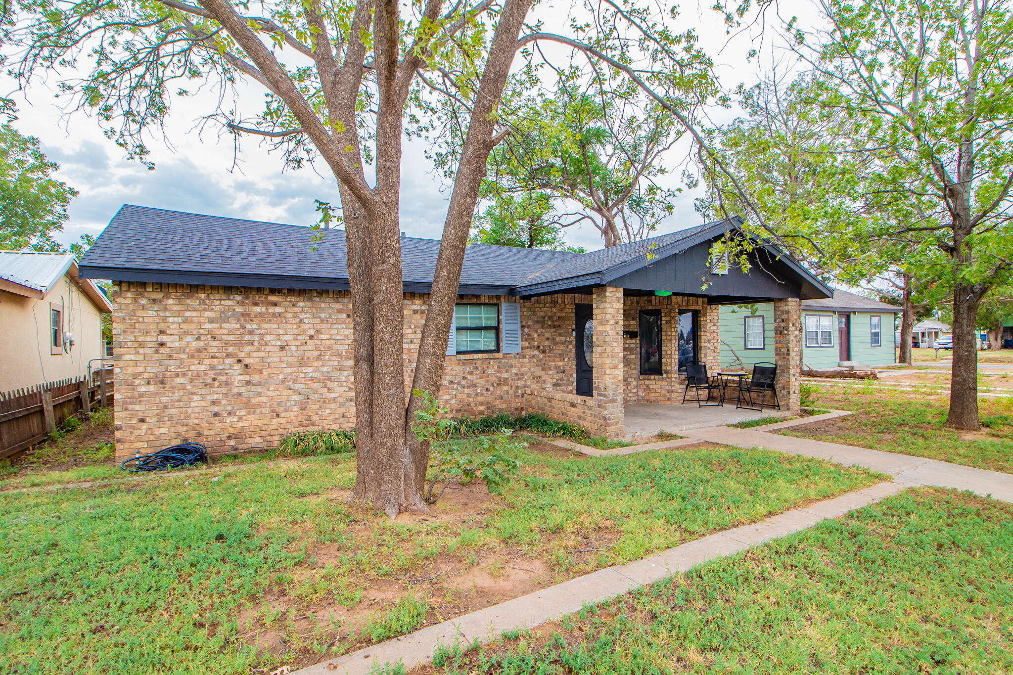 Single-story brick house with a covered porch, outdoor chairs, and mature trees in the front yard.