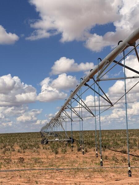 Center-pivot irrigation system spanning a dry, sparsely planted field beneath a cloudy blue sky.