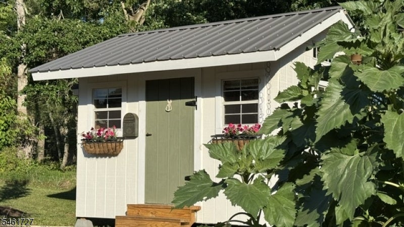 Cream garden shed with a dark metal roof, flower boxes, and wooden steps among leafy greenery.