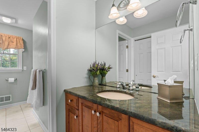 Bathroom with a wood vanity, dark stone countertop, large mirror, and window with a tan valance.