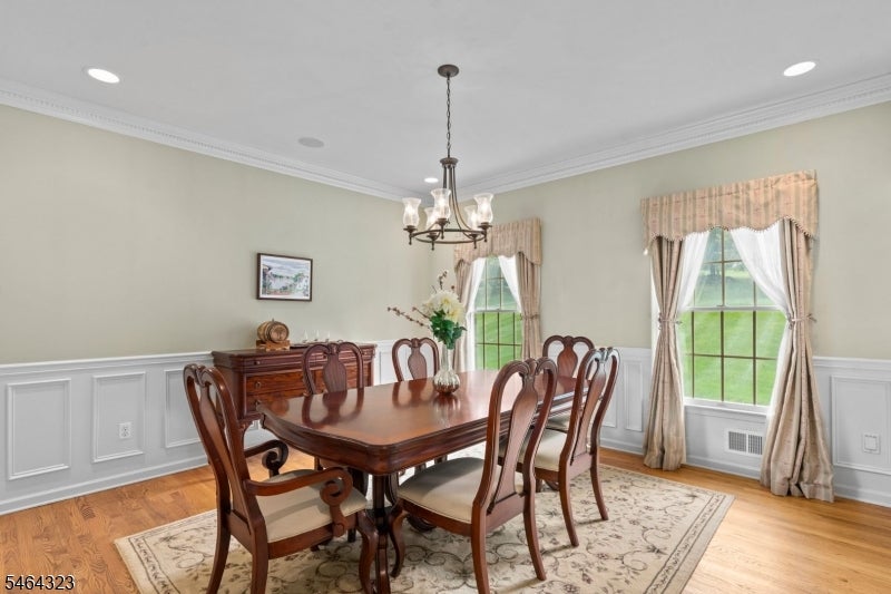 Furnished dining room with a wooden table, carved chairs, chandelier, and two curtained windows.