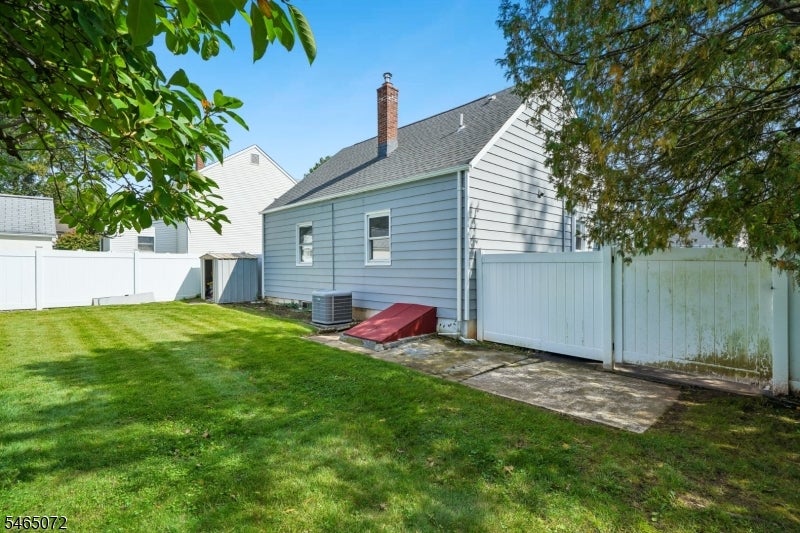 Side and rear of a blue-gray house beside a fenced lawn, with a paved patio and red basement access cover.
