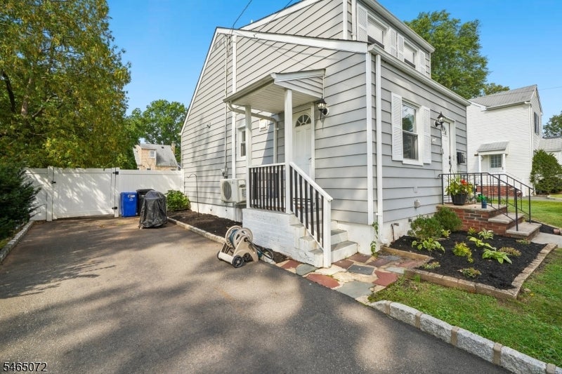 Light gray two-story house with a side entrance, paved driveway, white fencing, and landscaped garden beds.