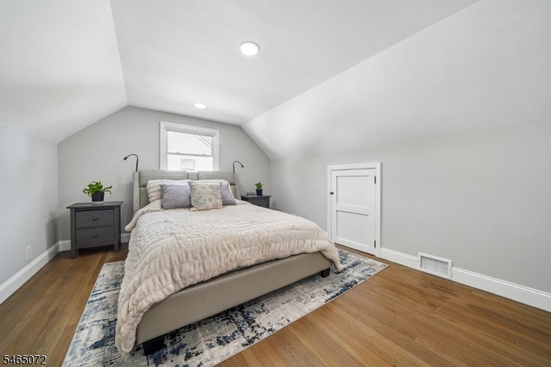 Furnished bedroom with a bed beneath a window, sloped ceiling, and hardwood floors.