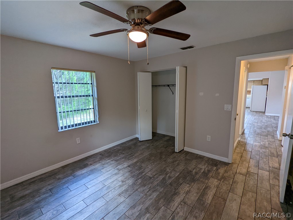 Empty bedroom with a ceiling fan, barred window, open closet, and dark tile flooring.
