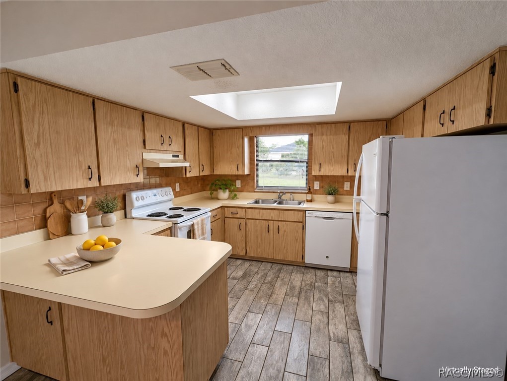 Kitchen with light wood cabinets, a breakfast peninsula, white appliances, and gray plank flooring.