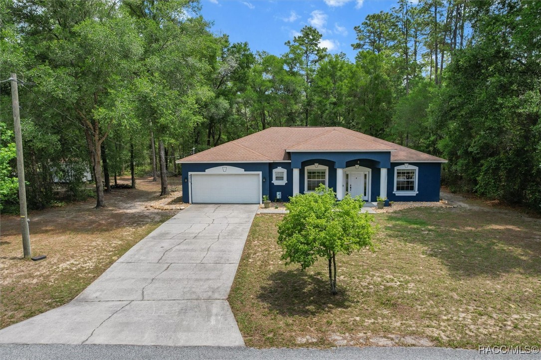Blue single-story house with white trim, an attached garage, and a long driveway on a wooded lot.
