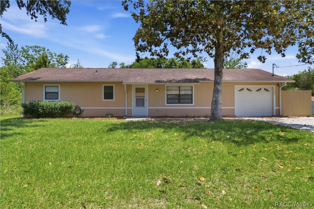 Single-story beige home with a white garage door, green lawn, and mature shade tree.