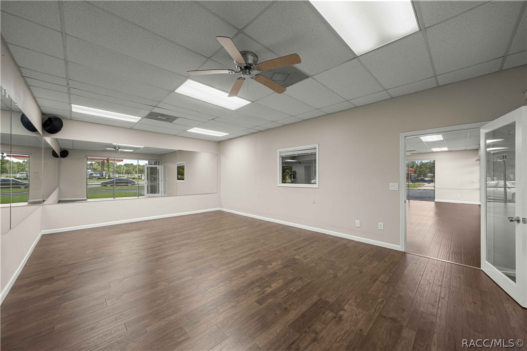 Vacant commercial room with wood-look flooring, mirrored wall, ceiling fan, and front windows.