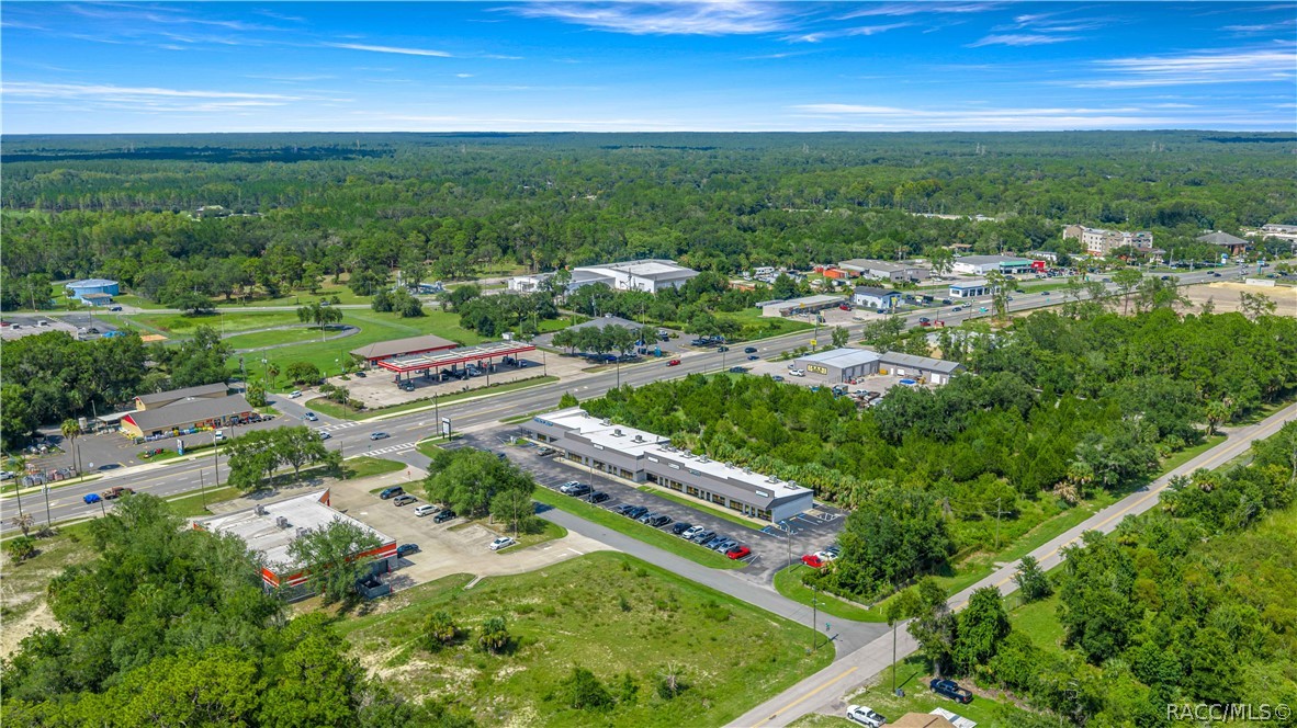 Aerial view of commercial buildings and parking lots along a road, bordered by dense woodland.