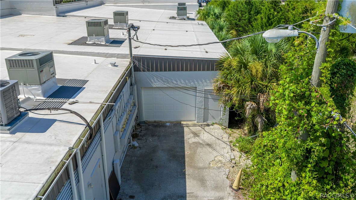 Building exterior with rooftop HVAC units, garage door, and concrete driveway beside dense greenery.