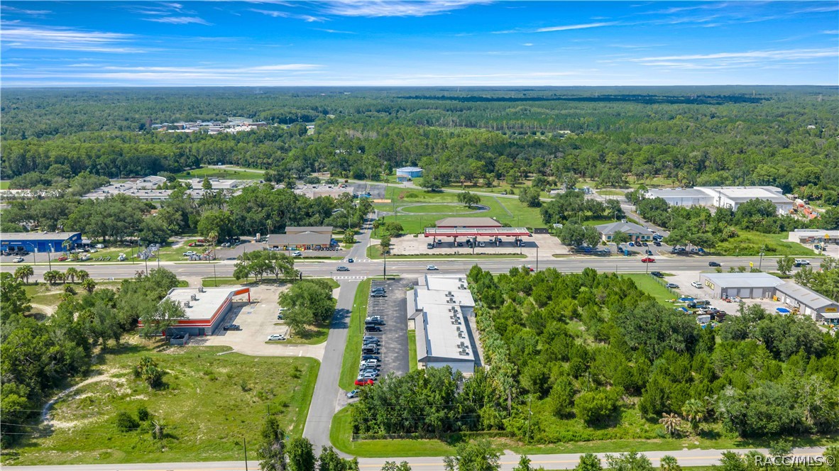 Aerial view of a commercial property and nearby businesses, roads, parking lots, and woodland.