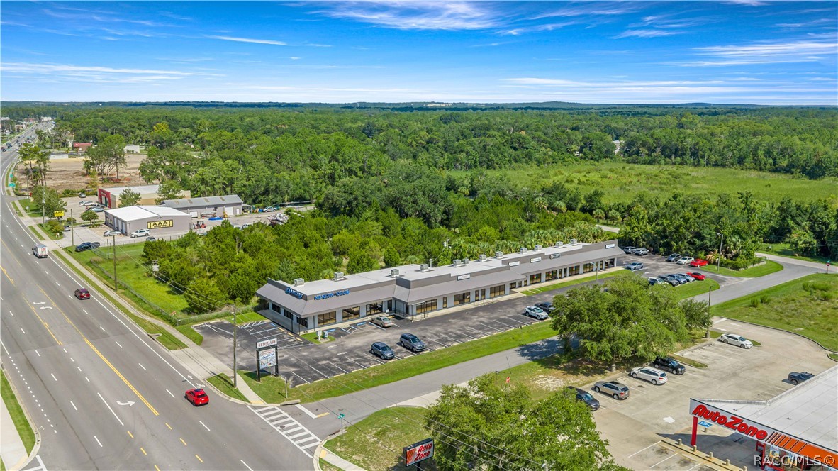 Aerial view of a commercial building and parking lot beside a multilane road, with wooded land behind it.