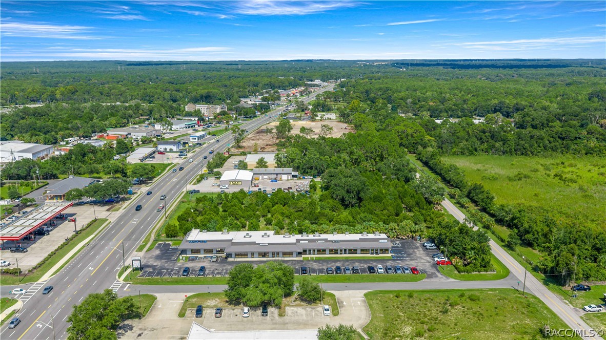 Aerial view of a commercial building and parking lot beside a major road, surrounded by trees and open land.