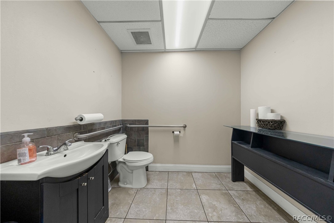 Bathroom with a white sink and toilet, tiled floor, grab bars, and dark shelving bench.