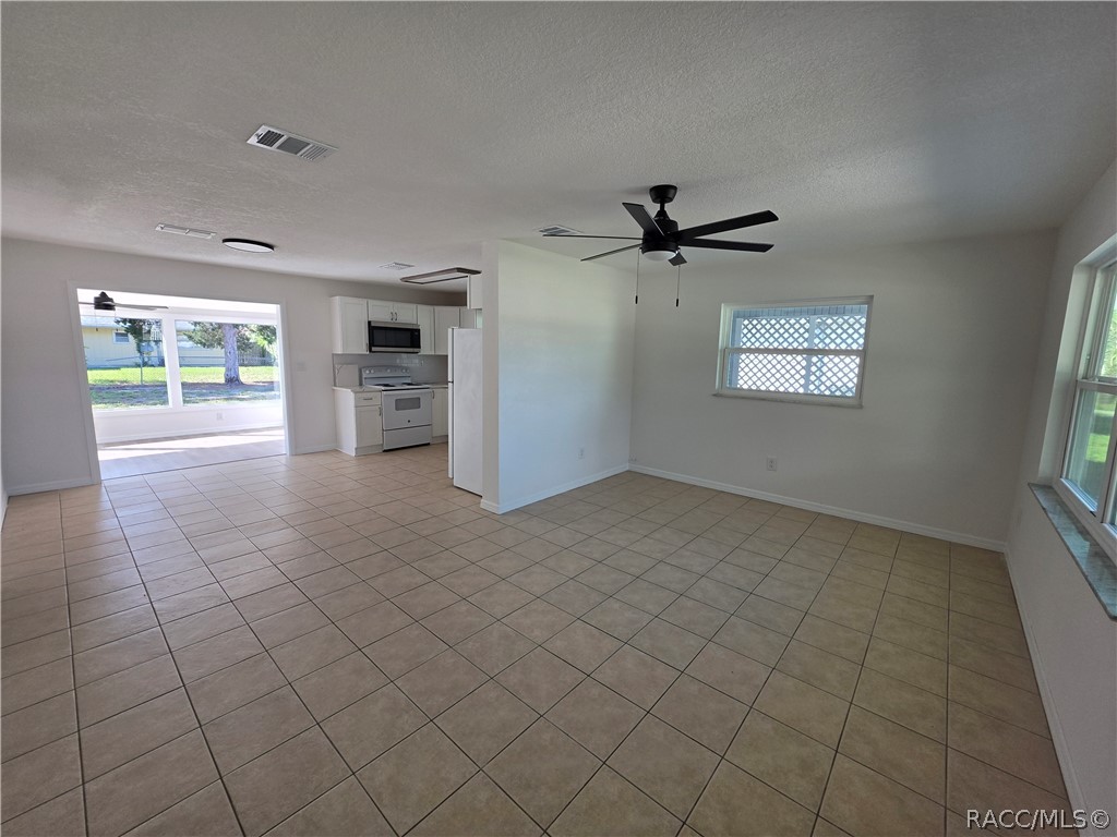 Empty tiled living area with a ceiling fan, large windows, and an open view into the kitchen.