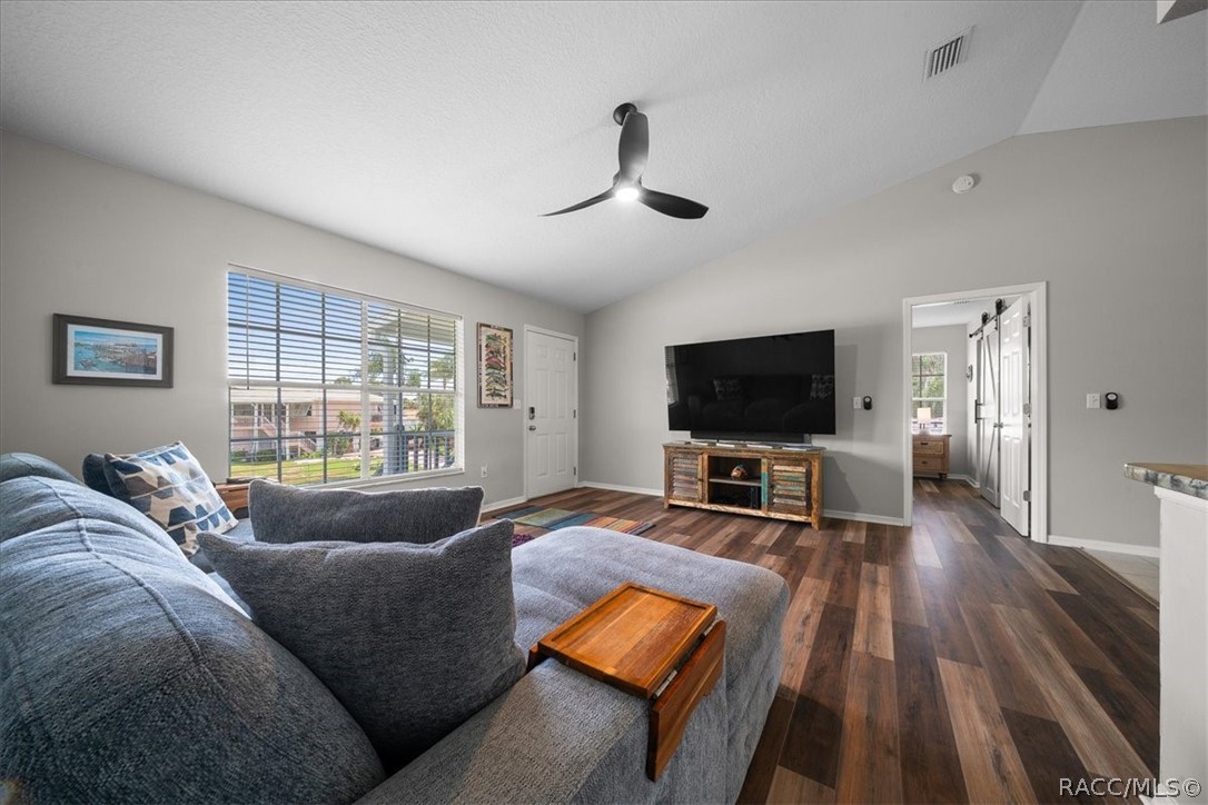 Living room with a gray sectional, wall-mounted television, ceiling fan, and wide window with blinds.