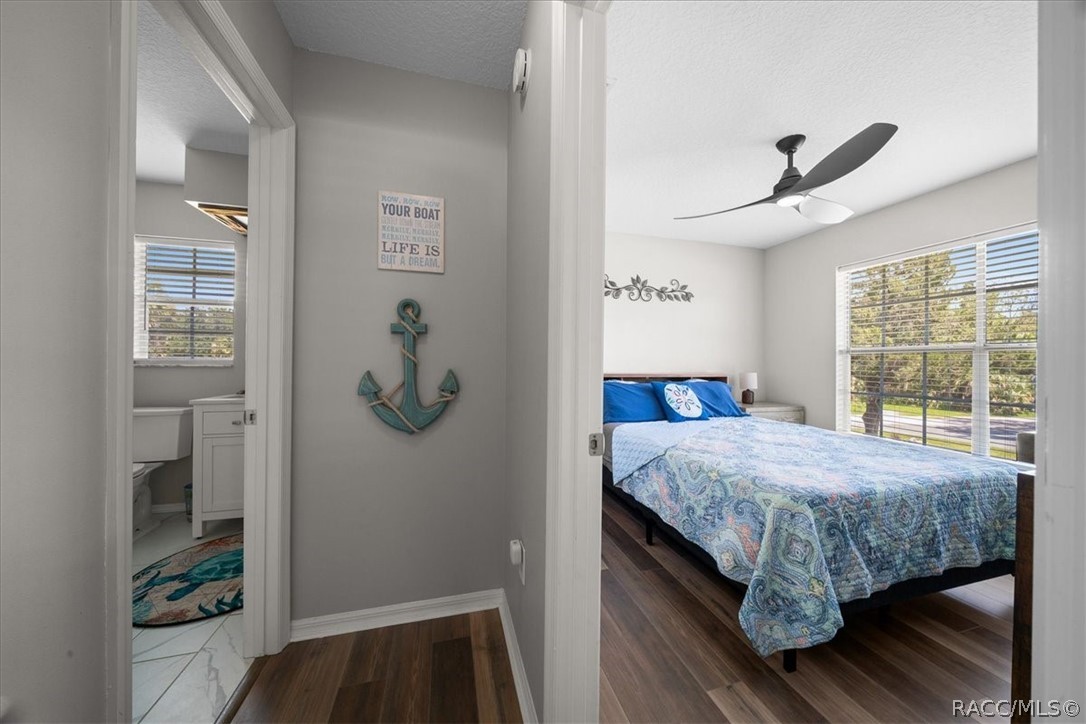 Bedroom with a blue patterned bedspread, ceiling fan, large window, and adjoining bathroom doorway.
