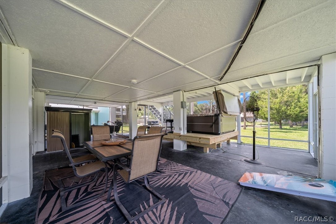 Covered patio with dining set, large grill, standing fan, and cornhole board overlooking a lawn.
