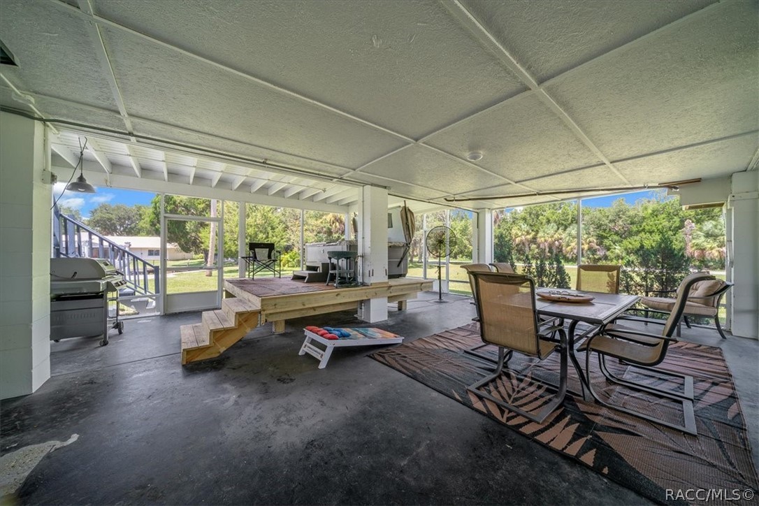 Covered patio with dining furniture, grill, and raised wooden platform overlooking a green yard.